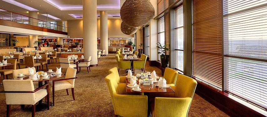 An expansive, sunlit dining area featuring stylish woven pendant lights, comfortable yellow armchairs, and large windows fitted with wooden blinds.