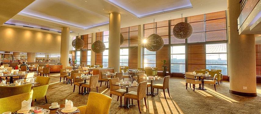 A spacious, sunlit dining area featuring floor-to-ceiling windows with wooden blinds, striking woven pendant lights, and neatly set tables ready for service.