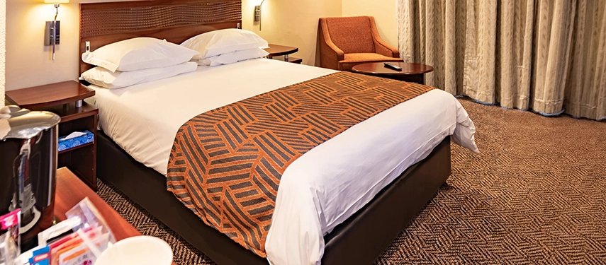 A comfortable, inviting guest room featuring a plush bed with a geometric runner, warm wood furnishings, and a dedicated workspace for the modern traveller.