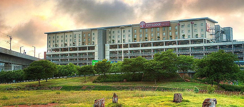 A wide, daytime view of the modern multi-story hotel, conveniently situated above a parking structure and framed by a tranquil stretch of green space.