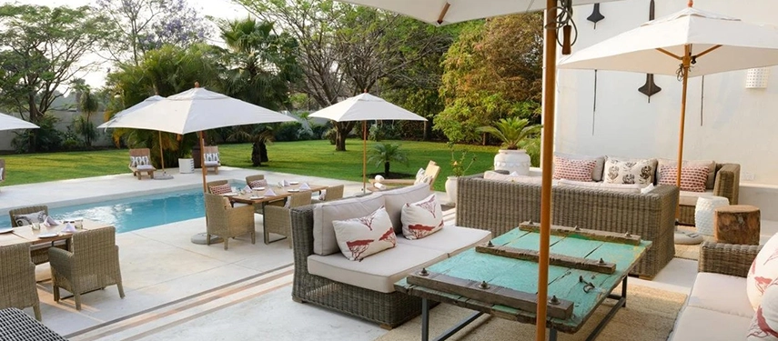 The poolside lounge terrace at Latitude 15° in Lusaka, with wicker sofas, printed cushions, a weathered turquoise coffee table and white parasols set against a lawn and mature tree canopy.