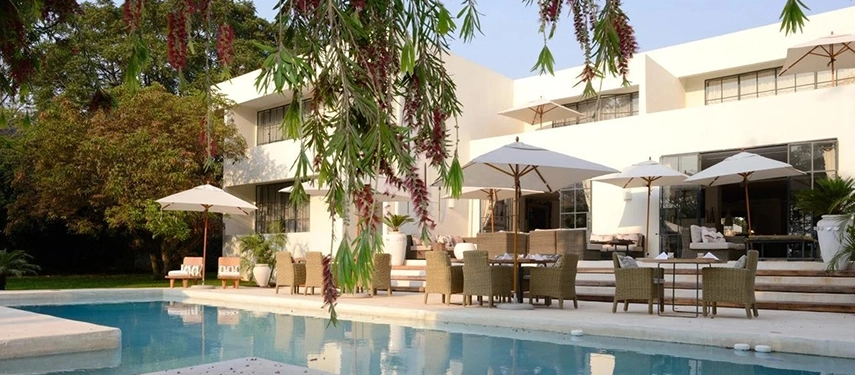 The swimming pool at Latitude 15° in Lusaka, with wicker dining chairs and white parasols arranged on the terrace, the hotel's white modernist facade rising behind a foreground of flowering tropical branches.