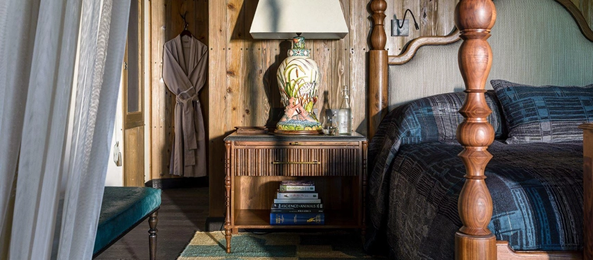 A close detail of a Xigera suite bedroom, showing the turned wooden post of a four-poster bed, a hand-painted ceramic lamp on a rattan bedside table, and a dressing gown hanging against timber-panelled walls.