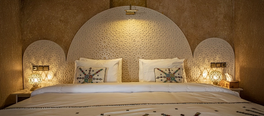 Elegant Moroccan bedroom with white carved headboard, soft lighting, and traditional embroidered cushions.