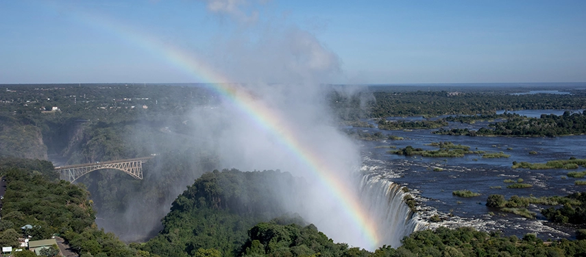 An aerial view of Victoria Falls shows mist rising from the gorge with a rainbow arching above the Zambezi River and the historic bridge in the distance.