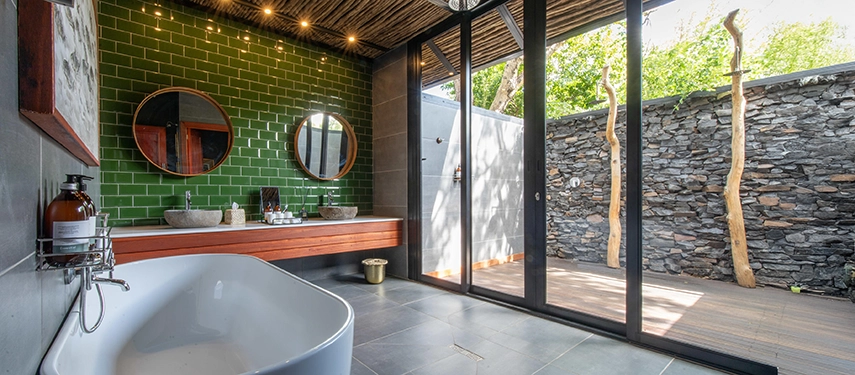 A contemporary bathroom at Mukwa River Lodge features a freestanding bath, twin basins, green tiled walls, and sliding doors opening onto a private outdoor shower courtyard.