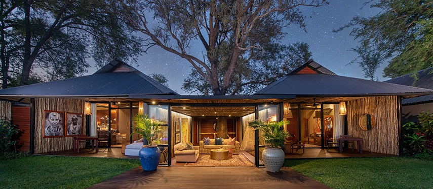 The main lodge at Mukwa River Lodge is softly illuminated at night, with open living spaces, elegant furnishings, and mature trees creating a warm, welcoming atmosphere.
