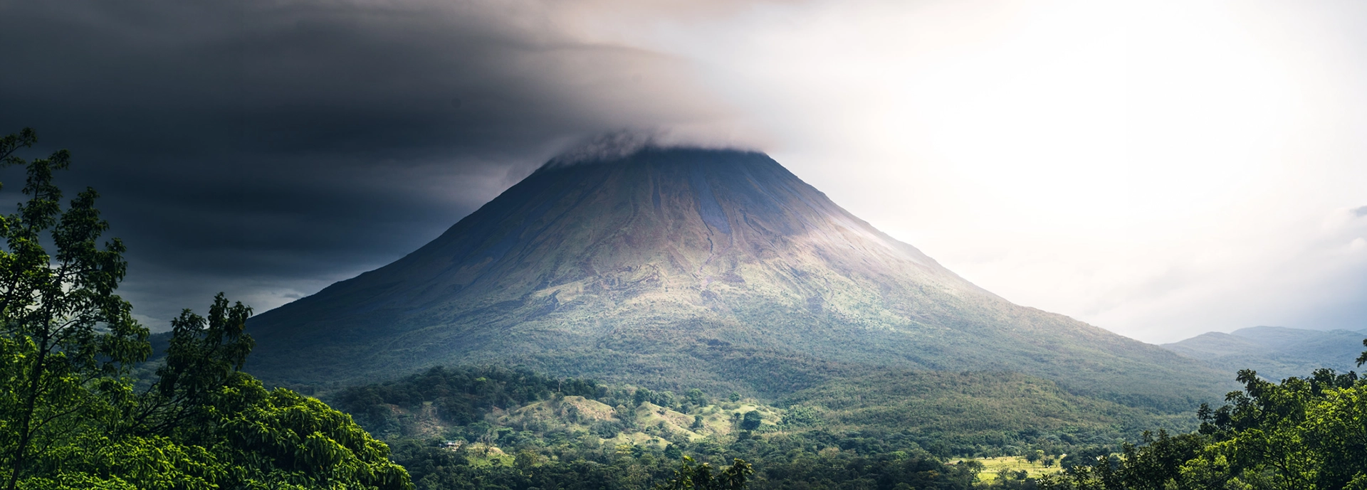 Arenal Volcano rising from the Costa Rican rainforest under a dramatic storm sky, its symmetrical cone partially shrouded in cloud with shafts of light breaking across the forested slopes below.