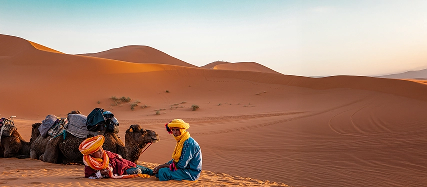 Two men in colourful Berber attire resting beside camels on a sand dune in the Moroccan desert.