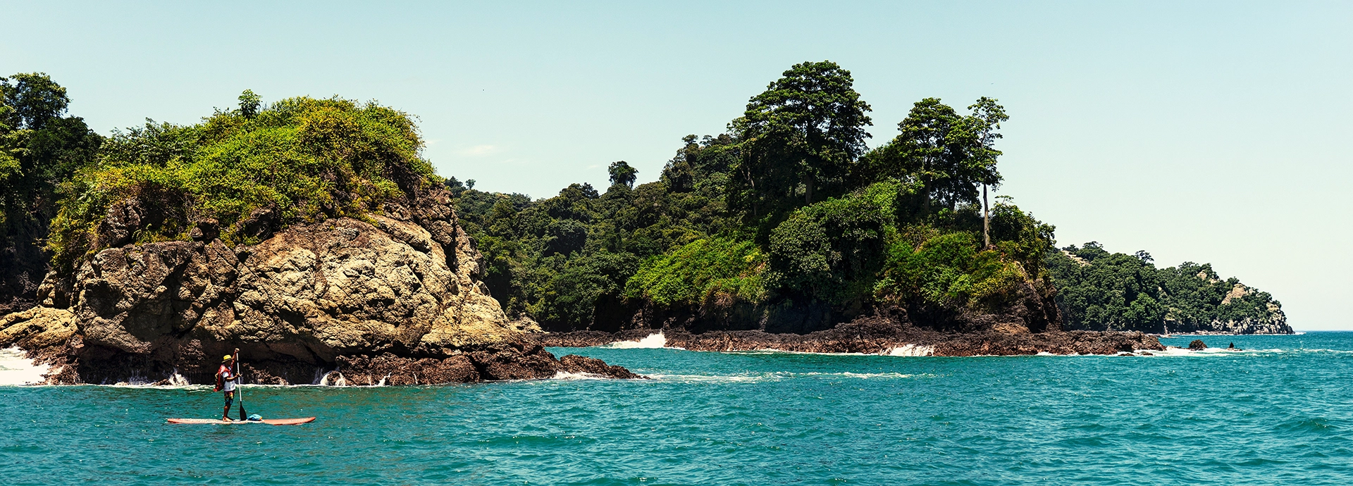 A stand-up paddleboarder navigates vivid turquoise waters alongside dramatic jungle-cloaked rock formations and headlands under a clear blue sky, Manuel Antonio, Costa Rica.