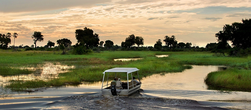 A small motorboat carrying a guide and guests heads into a winding Okavango Delta channel at sunset, the wide green floodplains glowing amber beneath a dramatic cloud-streaked sky.