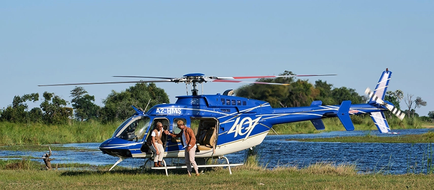 Two guests disembark from a blue helicopter on the edge of a Delta waterway, assisted by a guide, with vivid blue water and green reed banks stretching behind them under a cloudless sky.