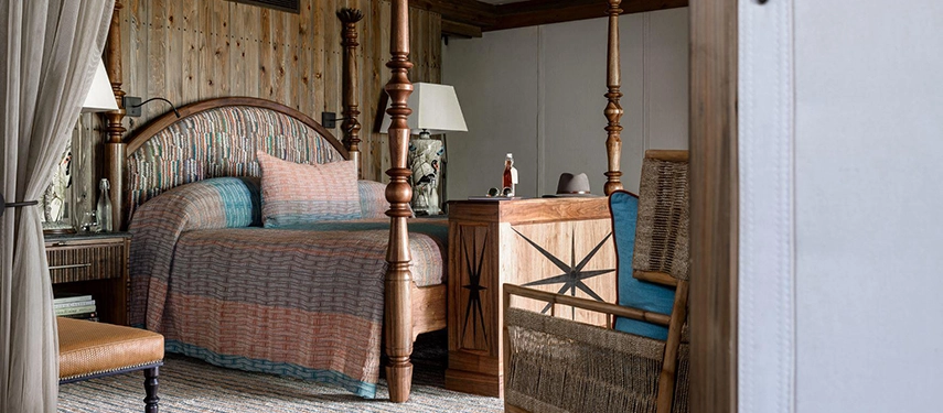 The four-poster bedroom of a Xigera suite, with turned wooden posts, a richly patterned headboard and bedspread in warm earth tones, a woven rattan armchair in teal, and reclaimed timber walls.