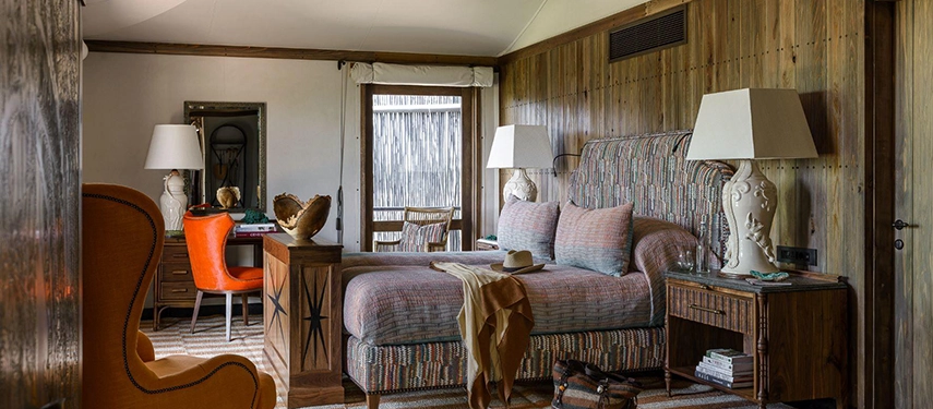 A Xigera suite sitting area and bedroom beyond, featuring a striped upholstered daybed, a vivid orange wingback chair at a writing desk, and timber-panelled walls dressed with warm lamplight.