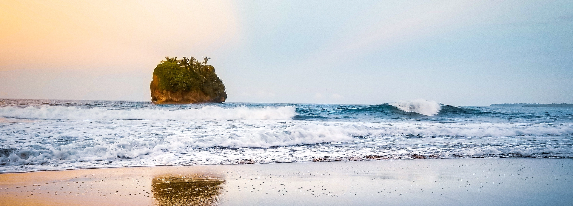 A small, vegetation-crowned rock island rises from the Pacific surf at dusk, golden light washing the sky above a wide, wave-kissed beach on Costa Rica's coastline.