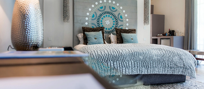 A close view of a guest room at Latitude 15° in Lusaka, with a textured grey bedspread, teal and brown cushions and a bespoke mandala motif painted across a wide timber headboard panel flanked by beaded wall hangings.